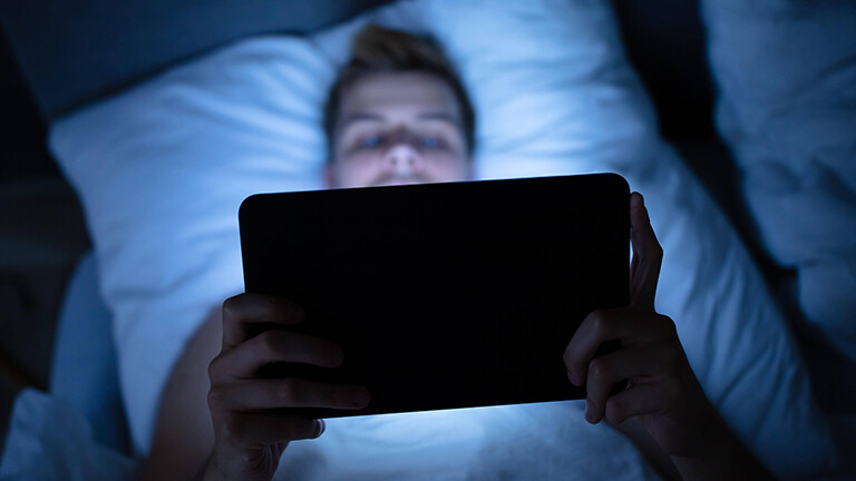 Foto eines Mannes, der auf dem Rücken im Bett liegt und auf einem Tablet einen Film schaut.