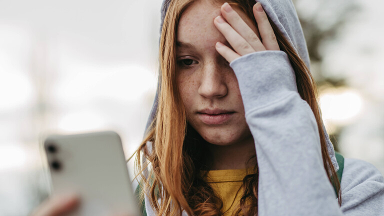 Foto einer jungen Frau, die traurig auf ihr Smartphone guckt und die Hand an die Stirn legt.