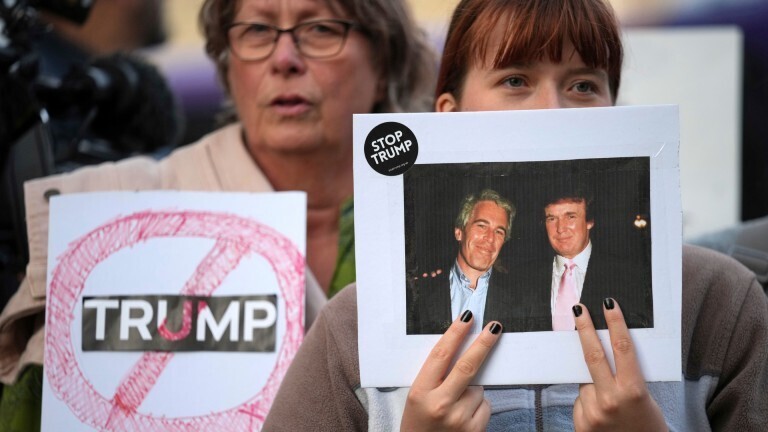 Bei einem Anti-Trump-Protest im englischen Windsor hält eine Frau ein altes Foto hoch, das Donald Trump und Jeffrey Epstein zusammen zeigt. Am Rand des Bildes klebt ein Sticker mit der Aufschrift "Stop Trump". Hinter der ersten Frau steht eine zweite Frau, die ein Plakat hochhält auf dem Trumps Name durchgestrichen ist.