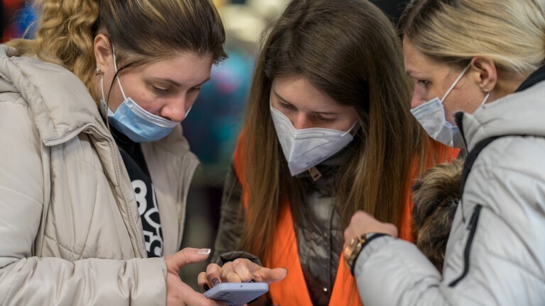 Das Foto zeigt eine Situation am Berliner Hauptbahnhof im März 2022. Zwei Frauen, die aus der Ukraine geflohen sind, bekommen Hilfe von einer Freiwilligen in einer orangefarbenen Weste. Alle Drei schauen gemeinsam auf ein Handy.