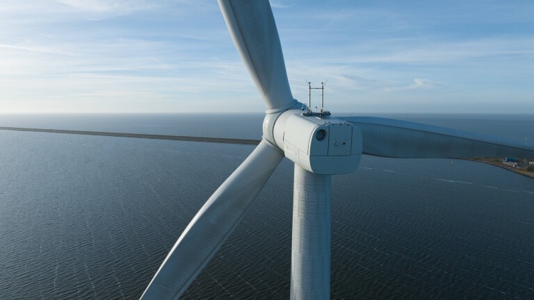 Das Bild zeigt eine Nahaufnahme der Rotorenblätter einer Windkraftanlage. Im Hintergrund ist Meer zu sehen und ein Teil der Küste.