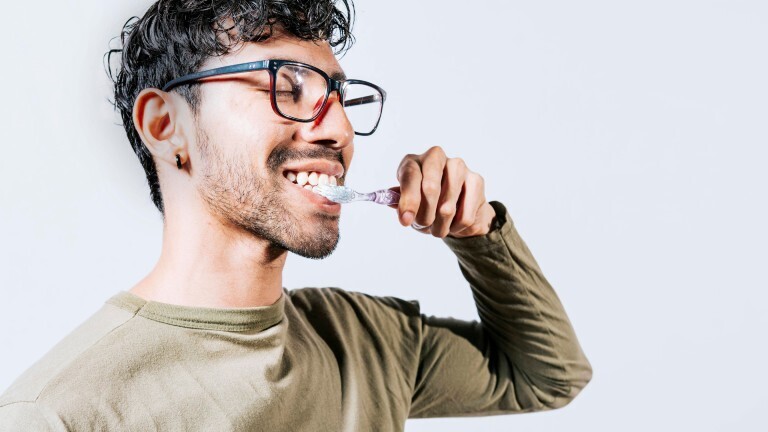 Ein Mann hält eine manuelle Zahnbürste an seine Zähne. Seine Augen sind dabei geschlossen. Er trägt eine Brille und wirkt jung.
