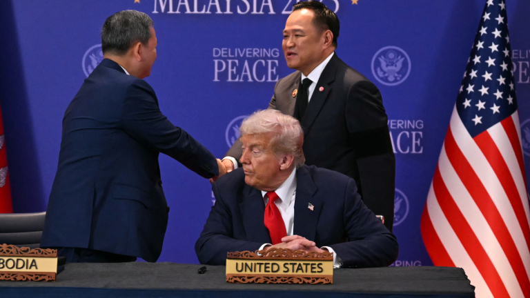 Zwei Männer in Anzügen schütteln sich stehend die Hände. Im Vordergrund sitzt ein weiterer Mann in Anzug und roter Krawatte vor einem Tischaufstellerchen. Darauf steht: United States.