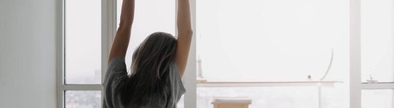 Eine Frau streckt sich, nachdem sie aufgewacht ist. Sie blickt aus den Fenstern aus ihrem Bett. Der Tag hat begonnen.