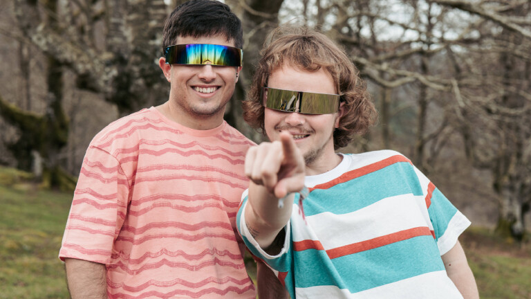 Zwei Männer mit flächigen Sonnenbrillen blicken in die Kamera. Einer der beiden zeigt direkt in deren Richtung.