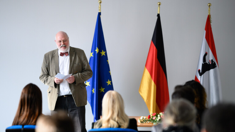 Ein Mann steht neben einer europäischen, einer deutschen und einer Berliner Flagge. Vor ihm sitzen mehrere Menschen auf Stühlen.