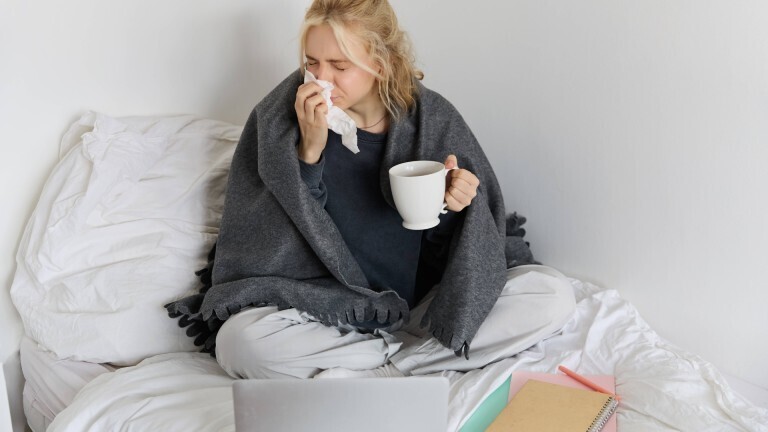 Eine Frau sitzt mit einer Decke um ihre Schultern auf einem Bett. Sie putzt sich mit einem Taschentuch die Nase und hält dabei eine Tasse in der Hand. Vor ihr auf dem Bett liegen ein Laptop und Notizbücher.