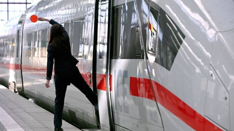 Eine Bahn-Mitarbeitende steht mit einem Fuß auf dem Gleis, mit dem anderen Fuß im ICE. Der Zug ist im Begriff, vom Hauptbahnhof München abzufahren.