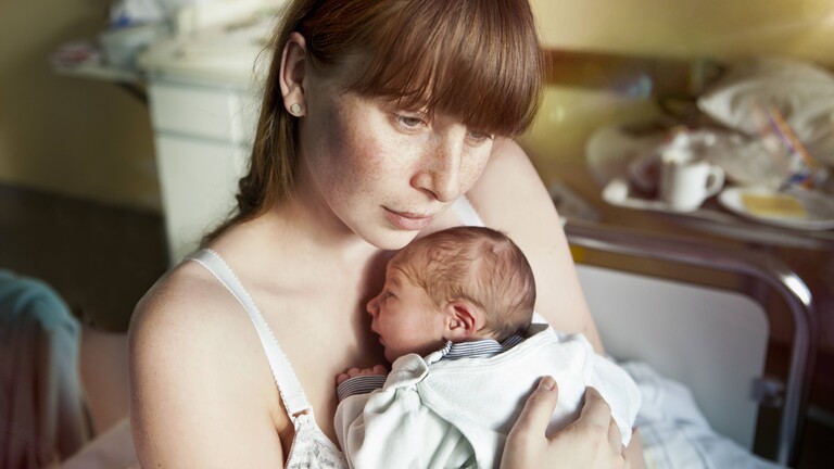 Eine junge Frau hält einen neugeborenen Säugling auf dem Arm und blickt traurig wirkend in die Leere.