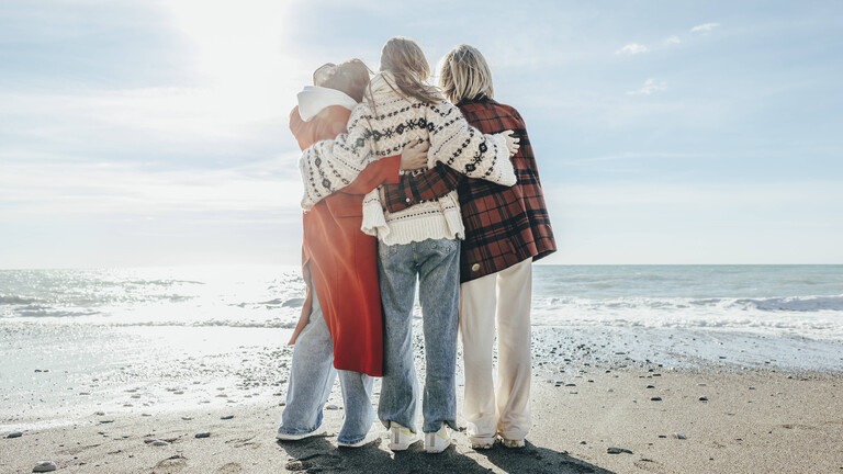Rückansicht von drei Frauen, die Arm in Arm am Strand stehen und aufs Meer gucken