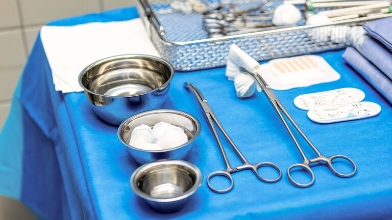 Ein detaillierter Blick auf einen sorgfältig vorbereiteten OP-Tisch in einem modernen Krankenhaus, reich an sterilen medizinischen Instrumenten. Das blaue Abdecktuch bildet den Hintergrund für eine Vielzahl von chirurgischen Werkzeugen, darunter glänzende Zangen, Scheren und Metallschalen, die akkurat angeordnet sind und für eine präzise Operation in der Chirurgie bereitliegen.
