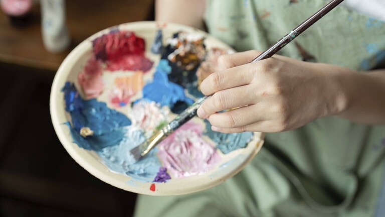 Hand einer Frau, die einen Pinsel in Farbe auf einer Farbpalette taucht.