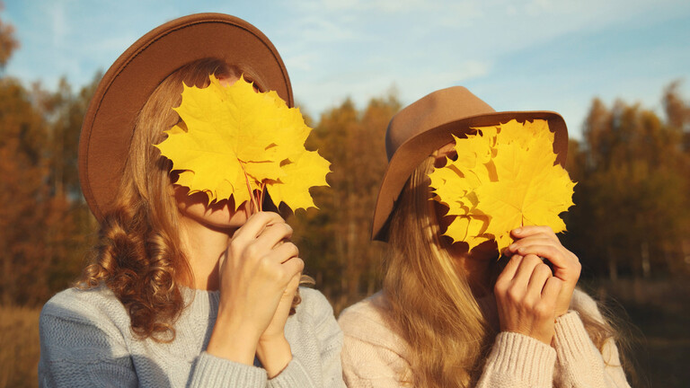 Zwei Frauen mit Hüten halten sich gelbe Blätter ins Gesicht