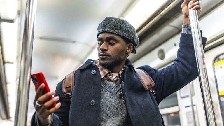 Ein Mann steht in einer U-Bahn. Er hält sich mit einer Hand an einer Stange fest. Mit der anderen Hand hält er sein Handy. Er schaut auf den Handy-Bildschirm.