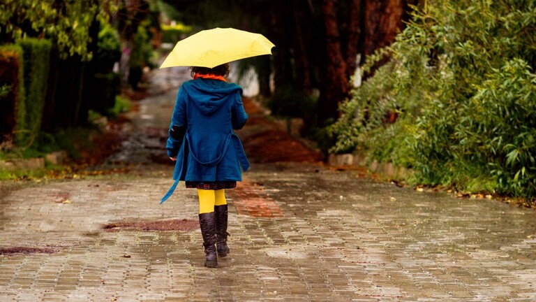 Rückenansicht einer Frau im Mantel, mit gelbem Regenschirm und Gummistiefeln.