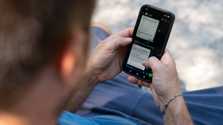 Ein Mann sitzt mit einem Smartphone im Freien und liest auf dem Display.