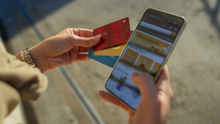 Nahaufnahme der Hände einer Frau, die im Freien ein Smartphone hält, auf dem eine Website eines Onlineshops geöffnet ist, und mehrere Kreditkarten