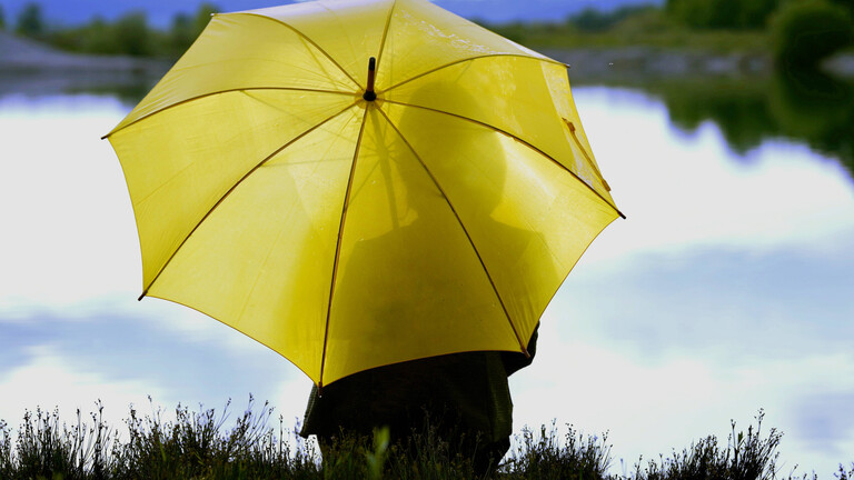 Auf einer Bank an einem See sitzt eine Frau. Sie hat einen großen gelben Regenschirm geöffnet.