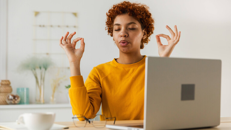 Eine Frau sitzt am Schreibtisch, hat die Augen geschlossen und Daumen und Zeigefinger berühren sich. Sie scheint zu meditieren.