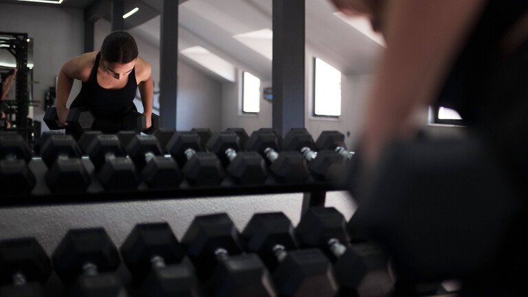 Eine Frau trainiert in einem Fitnessstudio mit Kurzhanteln. Sie sieht sich im Spiegel. Davor steht ein Regal mit weiteren Kurzhanteln.