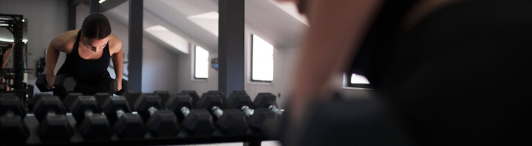 Eine Frau trainiert in einem Fitnessstudio mit Kurzhanteln. Sie sieht sich im Spiegel. Davor steht ein Regal mit weiteren Kurzhanteln.