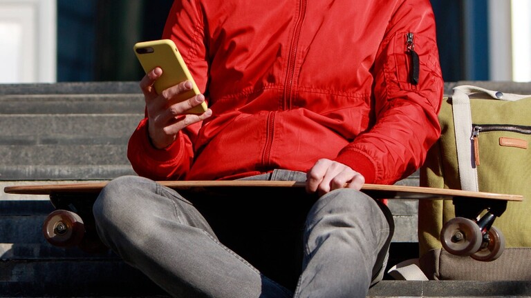 Mann auf einem Longboard, der sein Smartphone benutzt, auf einer Treppe sitzt und mit seinem Longboard und Skateboard im Freien sitzt.
