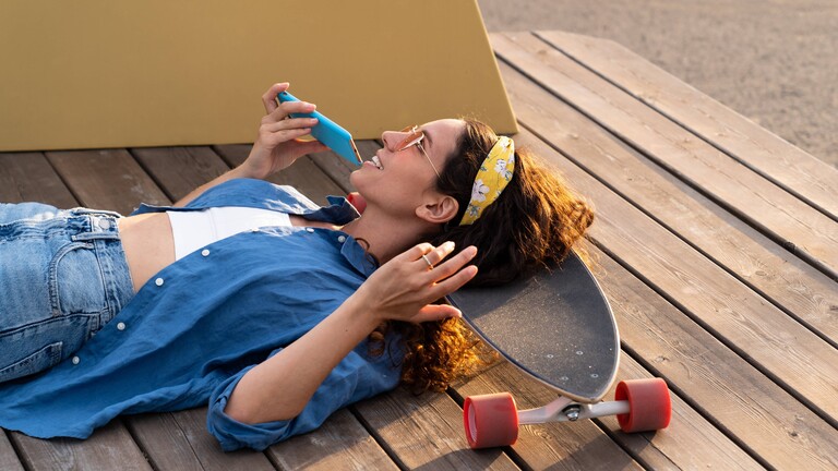 Eine frau liegt mit dem Kopf auf einem Skateboard und spricht eine Sprachnachricht in ihr Smartphone.