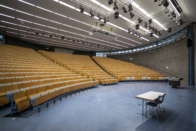 Blick in das leere Audimax der Technischen Universität Dortmund. Viele Stuhlreihen mit klappbaren Pulten links, rechts im Bild steht ein Tisch mit zwei Stühlen.