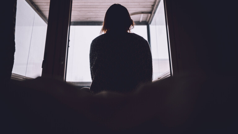 Eine Frau sitzt auf einem Bett und schaut aus dem Fenster. Wir sehen sie von hinten.