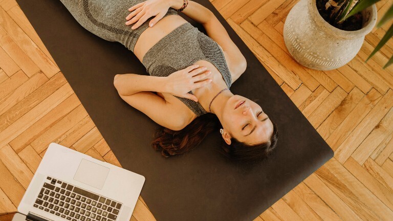 Eine Frau liegt auf dem Rücken auf einer Matte. Sie trägt Sportkleidung und hat ihre Hände jeweils auf die Brust und den Bauch gelegt.