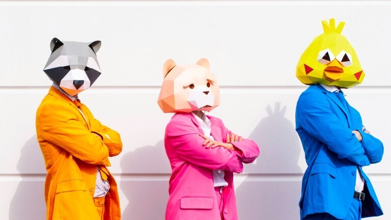 Drei Personen in bunten Anzügen und Tiermasken posieren nebeneinander vor einer weißen Wand.
