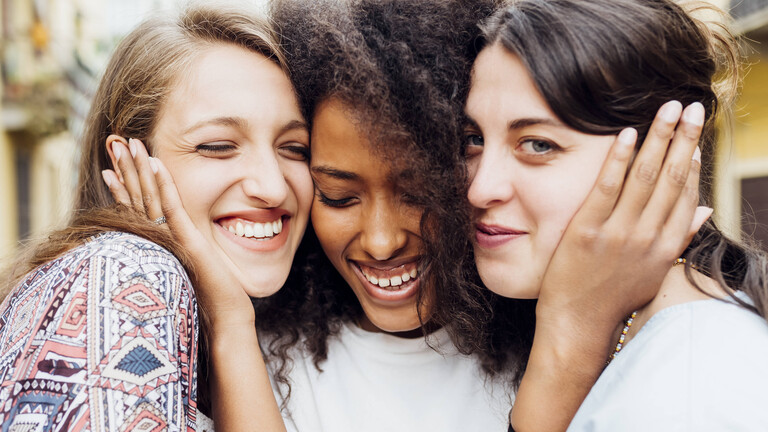 Drei Frauen halten ihre Gesicht innig nebeneinander. Die Frau in der Mitte hält ihre Hände an das Gesicht der beiden anderen Frauen. Sie lachen.