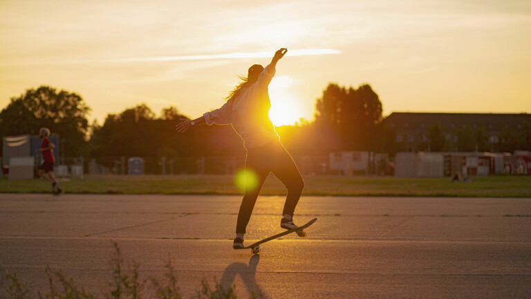 Eine junge Frau, fährt bei Sonnenuntergang mit ihrem Skateboard auf dem Tempelhofer Feld.
