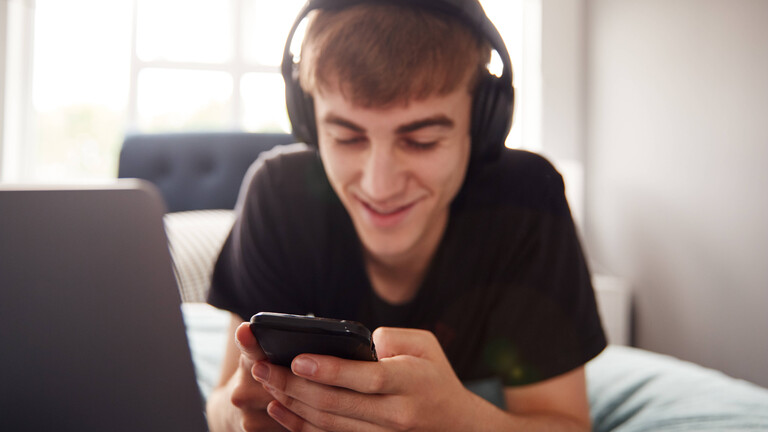 Ein junger Mann mit Kopfhörern auf dem Kopf liegt auf dem Bett vor einem aufgeklappten Laptop und schaut auf sein Smartphone.