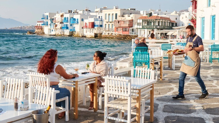 In einem Restaurant auf Mykonos, das direkt am Meer liegt, bringt ein Kellner Essen zu Gästen, die am Tisch sitzen.