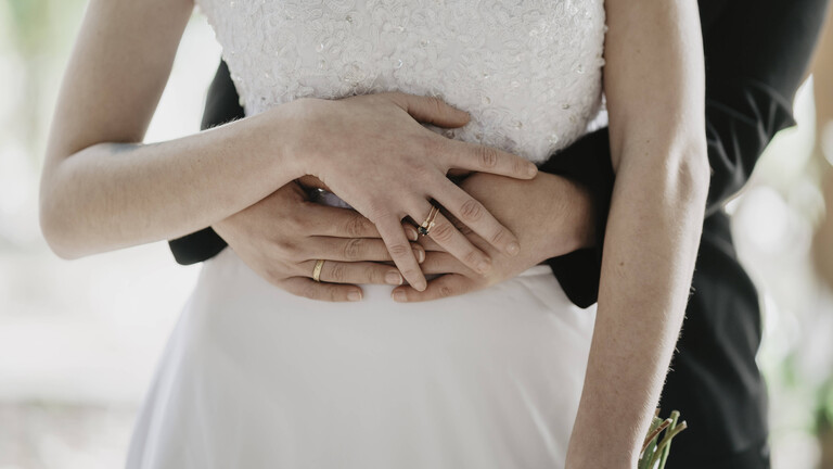 Ein Hochzeitspaar steht hintereinander. Eine der beiden Frauen trägt ein weißes Hochzeitskleid. Die andere Frau trägt einen schwarzen Anzug. Sie umarmen sich, dabei halten sie ihre Hände übereinander.