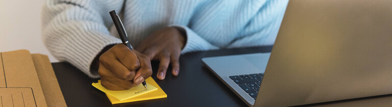 Frau schreibt etwas auf ein Postit, neben ihr steht ein Laptop