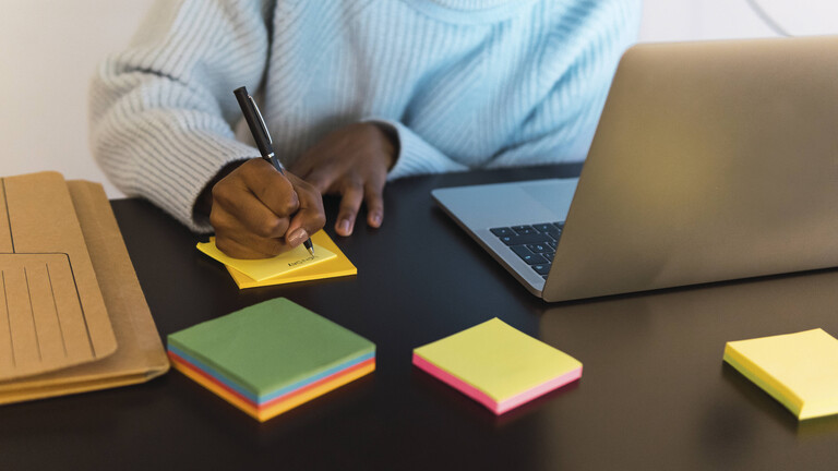 Frau schreibt etwas auf ein Postit, neben ihr steht ein Laptop