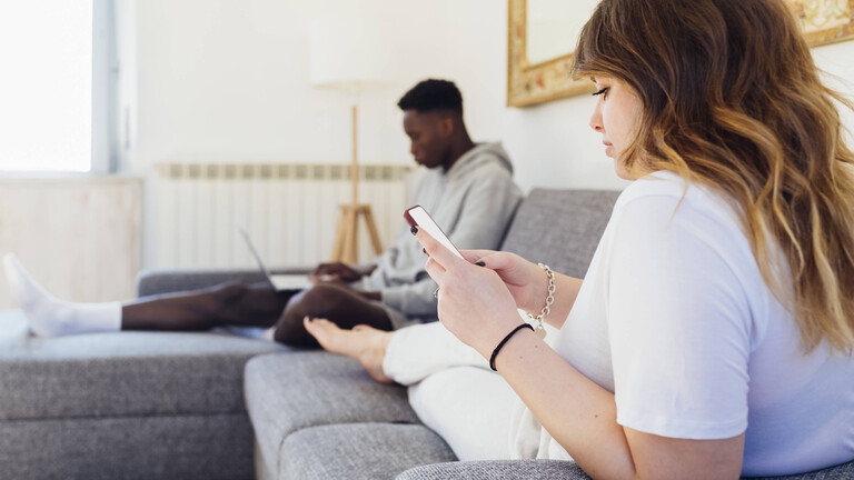 Ein junger Mann und eine junge Frau sitzen gemeinsam auf einer Couch. Er schaut auf seinen Laptop und sie schaut auf ihr Handy.