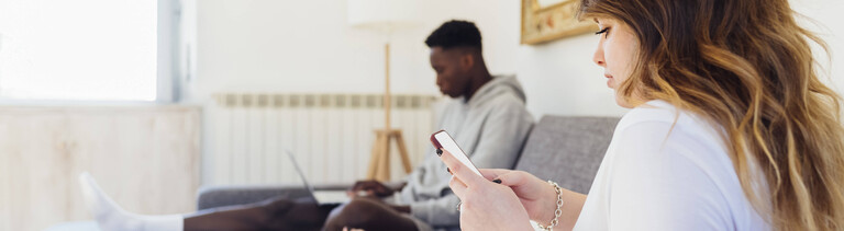 Ein junger Mann und eine junge Frau sitzen gemeinsam auf einer Couch. Er schaut auf seinen Laptop und sie schaut auf ihr Handy.