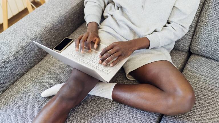 Person sitzt mit Laptop auf den Beinen auf einem Sofa, daneben liegt ein Smartphone