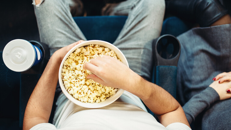 Eine männliche Person sitzt in einem Kinosessel und greift in einen Eimer mit Popcorn