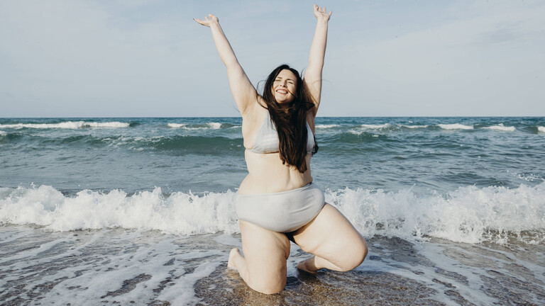 Eine Frau im Bikini kniet an einem Strand im Wasser und hebt ihre Arme in die Luft. Sie lächelt und hat ihre Augen geschlossen.