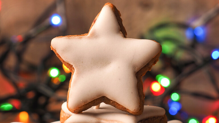 Auf einem Tisch sind Zimtsterne aufeinandergestapelt. Dahinter liegt eine Lichterkette mit bunten Lichtern.