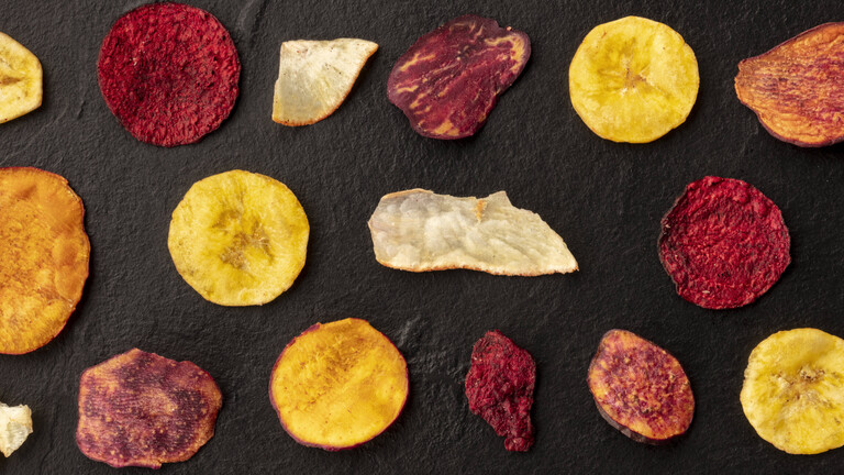 Verschiedene Chipssorten, unter anderem auch Gemüsechips, liegen nebeneinander aufgereiht auf einem schwarzen Untergrund.