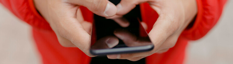 Foto der Hände eines jungen Mannes, der eine rote Daunenjacke trägt und mit beiden Daumen auf einem Smartphone tippt