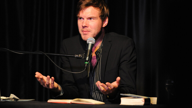 Autor und Musiker Thorsten Nagelschmidt sitzt bei einer Lesung an einem Tisch. Vor ihm liegt ein aufgeklapptes Buch. Er spricht in ein Mikrofon.