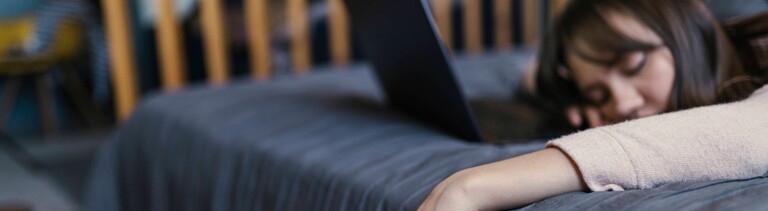 Eine Frau liegt auf dem Bett, die Hand, in der sie ein Smartphone hält, hängt schlaff herab, Vor ihr ein Laptop.