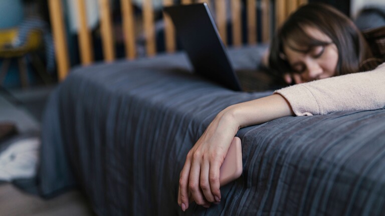 Eine Frau liegt auf dem Bett, die Hand, in der sie ein Smartphone hält, hängt schlaff herab, Vor ihr ein Laptop.