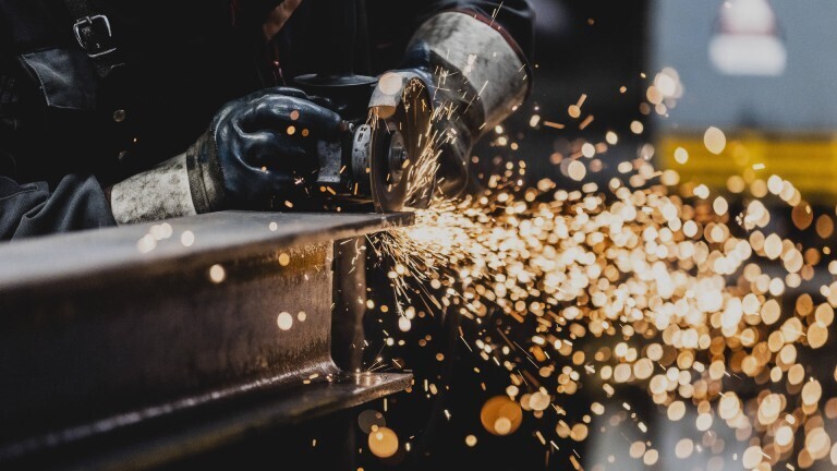 Ein Metallbauer bearbeitet einen Stahlträger mit einem Trennschleifer. Dabei sprühen helle Funken.